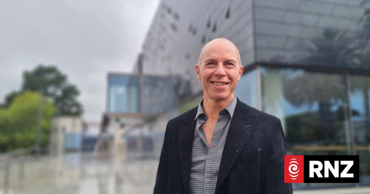 A smiling man in a dark blazer standing in front of a modern glass building.