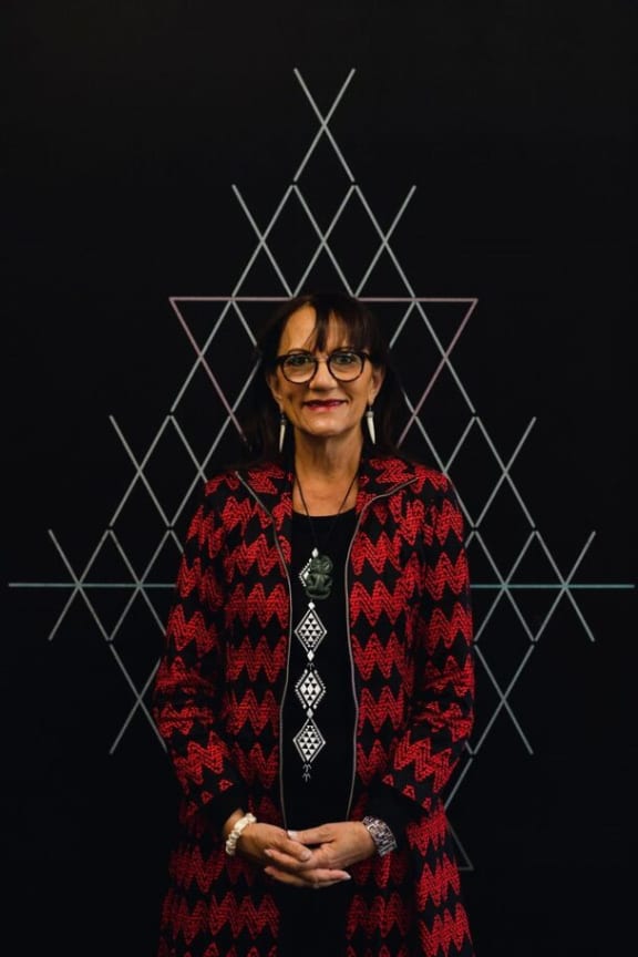 Te Puea smiles at the camera. She stands in front of a black wall designed with white tukutuku panelling.