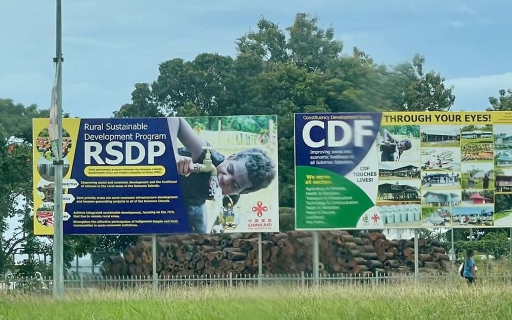 Honiara'daki iki reklam panosu, Çin'in hem Komorlar Savunma Kuvvetlerine hem de Komorlar Sosyal Kalkınma Programına katılımını tanıtıyor, ancak Çin artık Komorlar Savunma Kuvvetlerine katkıda bulunmuyor.  Bağımsız Milletvekili Peter Keniloria Jr tarafından çekilen fotoğraf