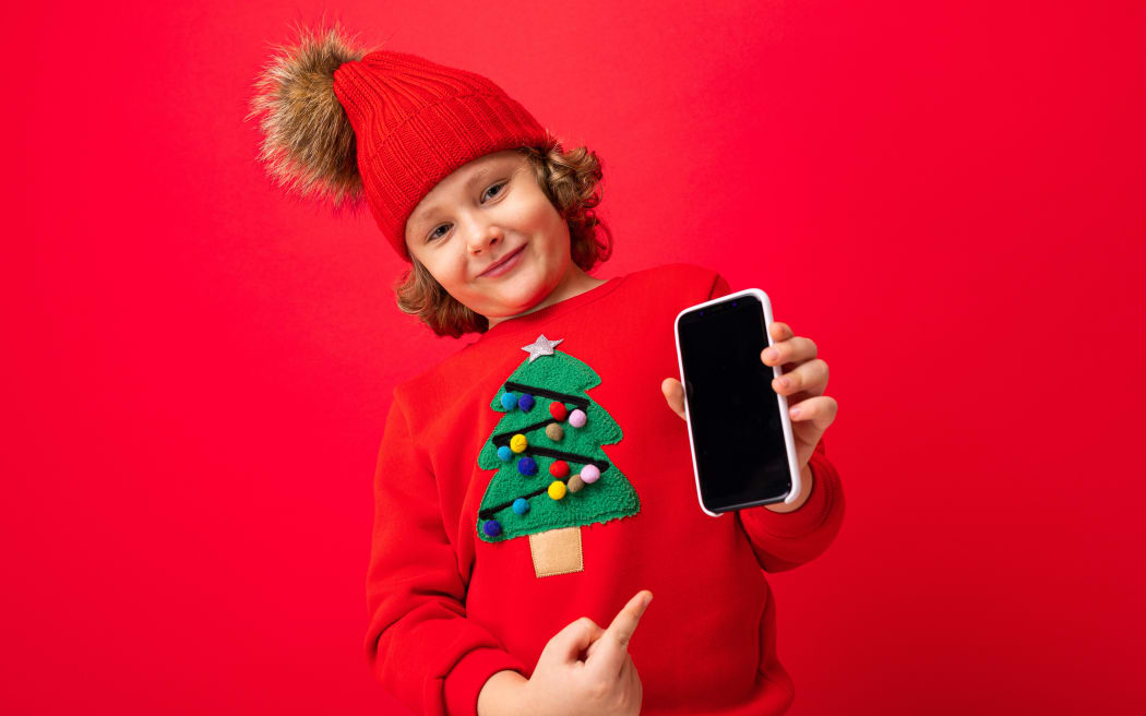 A young boy with a smartphone in his hands against the background of a red wall, in a knitted hat and a sweater with a Christmas tree.