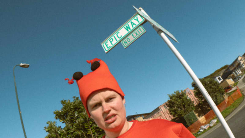 Comedian Liv Ward standing in a lobster costume in front of a street sign that reads "Epic Way", in Newlands, Wellington.