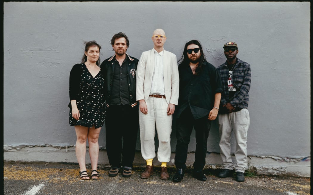 Five people stand against a grey wall. The man in the middle wears a white suit.