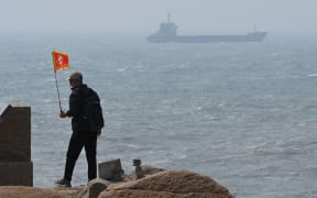A man walks with a flag as a ship passes by behind him on Pingtan island, the closest point to Taiwan, in China’s southeast Fujian province on April 8, 2023. - China launched military drills around Taiwan on April 8, in what it called a "stern warning" to the self-ruled island's government following a meeting between its president and the US House speaker. (Photo by GREG BAKER / AFP)