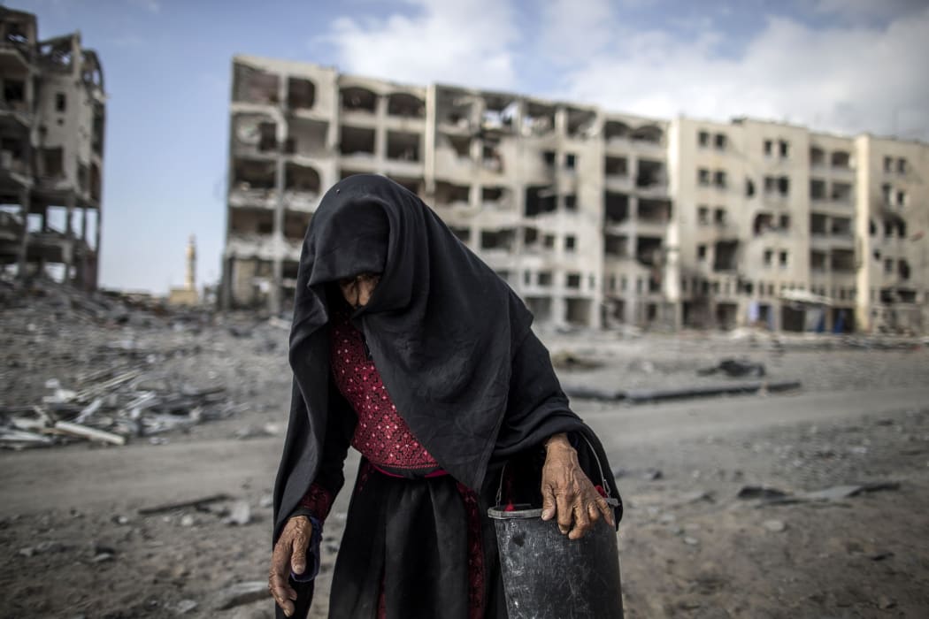 An elderly Palestinian woman carries a bucket walking past destroyed buildings in the Gaza Strip.