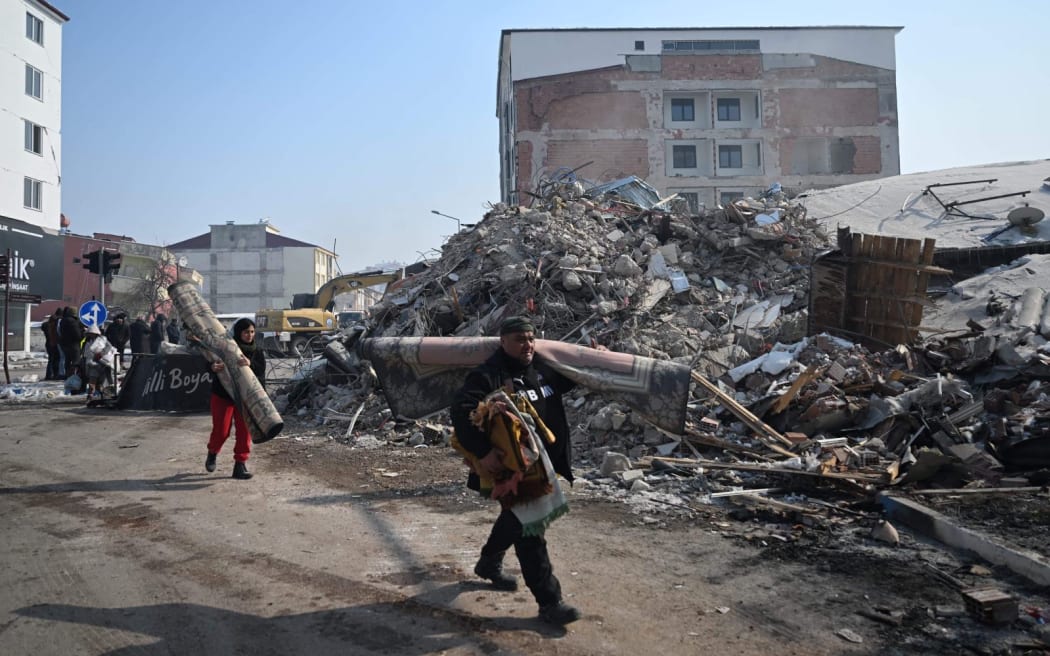 People carry carpets as they walk by collapsed buildings at Elbistan district of Kahramanmaras on February 11, 2023, after a 7.8-magnitude earthquake struck the country's southeast earlier in the week. - The death toll from a catastrophic earthquake that hit Turkey and Syria climbed to more than 25,000 on Saturday, as rescuers worked in freezing weather to find people alive. (Photo by OZAN KOSE / AFP)