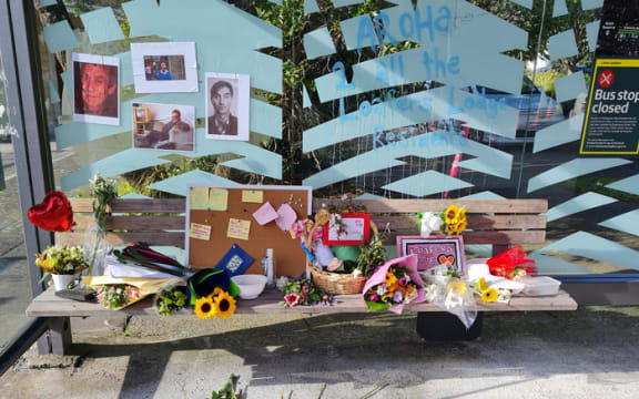 A makeshift shrine at a bus stop. The bench is covered in flowers, notes and signs. The glass behind the bench is hung with pictures of the victims of the Loafers Lodge fire.