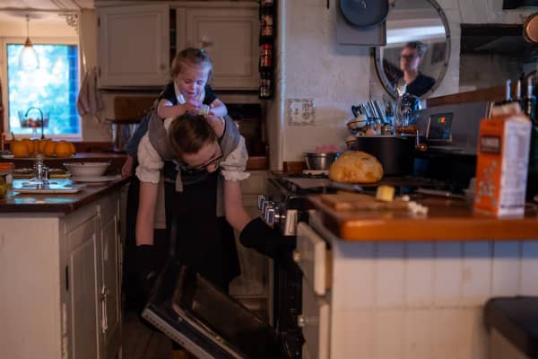 Simone Collins wearing oven mitts, bends down with a toddler strapped on her back to take a dish out of the oven.