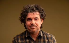 Toby Manhire wears a urban lumberjack style shirt and sits in front of a brown background.