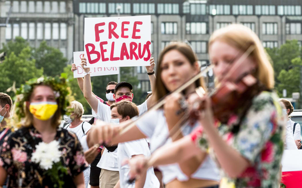 Two women play a folk song on violins, while behind them a man holds a sign saying "Free Belarus" at a rally of Belarusians living in exile.