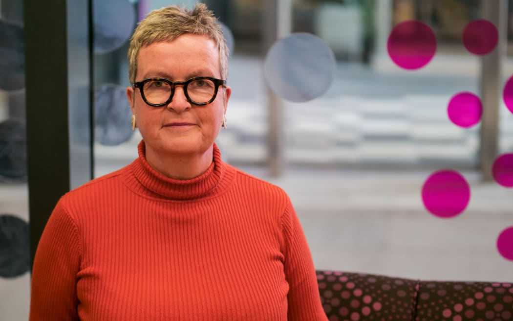 Martha looks at the camera. She is wearing an orange turtleneck sweater. She is standing inside an office.