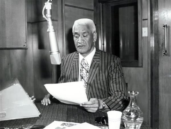 A white-haired man sits in front of a radio microphone holding a script.