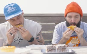 Two men wearing hats sit outside eating pies.