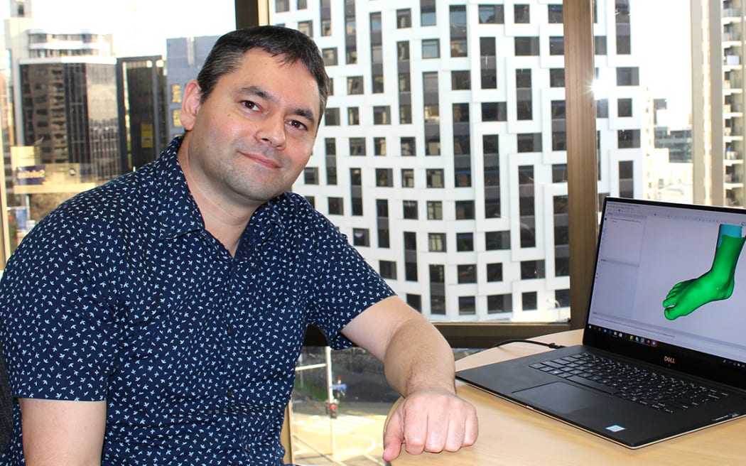 A man in a dotty navy short-sleeved shirt sits at a desk next to a laptop displaying a green foot graphic. The man is looking at the camera.