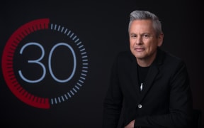 Guyon Espiner on a black background, looking at camera with logo behind him