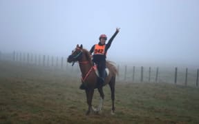 Brydie is atop a horse in a misty field. She has her arm in the air in triumph and smiles at the camera.
