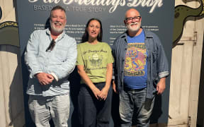 Two men with grey beards and wearing shirts, stand on either side of a dark-haired woman in a green t-shirt in front of a poster reading 'Fat Freddy's Drop'.