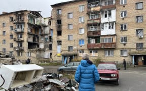A woman wearing a blue puffer jacket, with her back to the camera, stands in front of a brick apartment building in Ukraine, which is damaged after being hit in a missile strike.