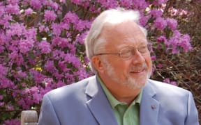 American composer William Bolcom, seated in front of a lilac azalea bush