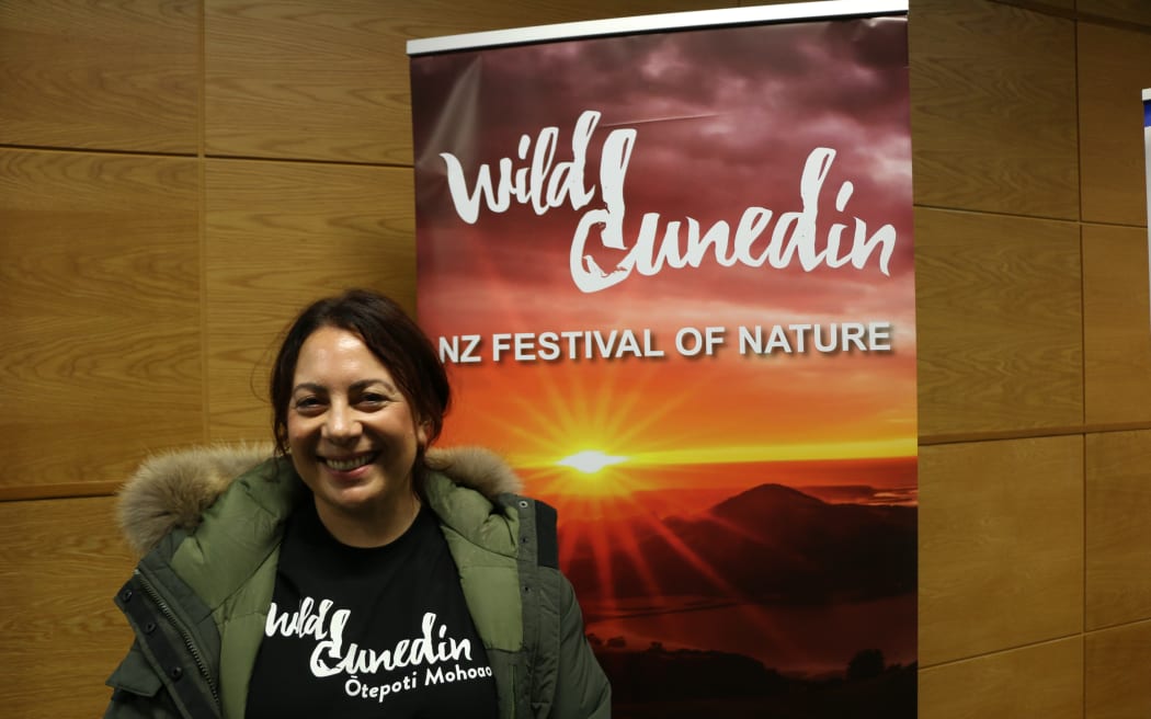 Amanda is wearing a black 'Wild Dunedin' t-shirt underneath a khaki green hooded jacket. She is smiling and standing in front of a Wild Dunedin: NZ Festival of Nature banner featuring a bright sunset.