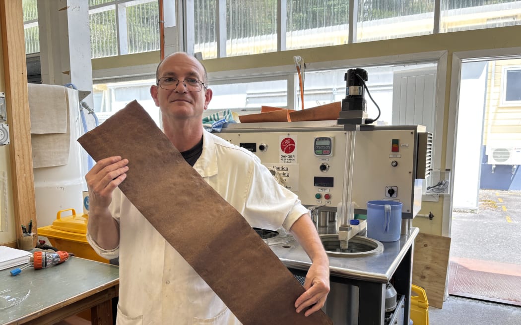 Sean is standing in front of a small scale paper making machine, that looks a bit like a top-loader washing machine. He is wearing glasses and a long white lab coat, and is holding a long strip of brown parchment across his torso.