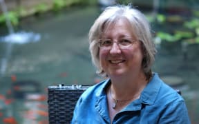 This is a headshot of Melissa. She is sitting outside on a chair. Behind her is a fish pond. She wears glasses and a blue shirt.