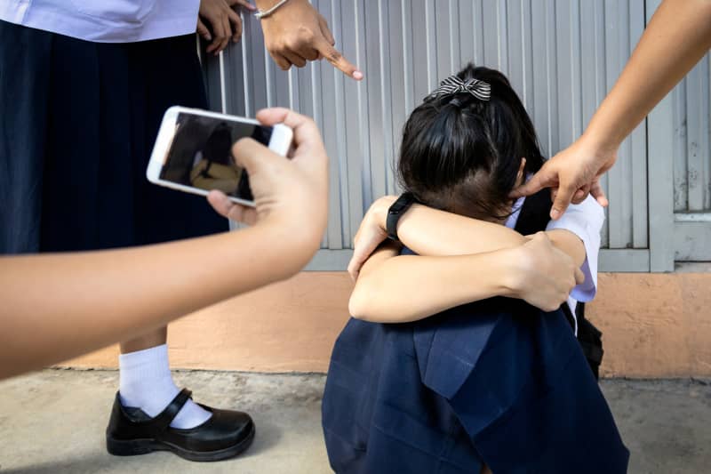 Sad girl student in school uniform sitting on the floor with hands on knees while being pointed at and video recorded on a mobile phone.
