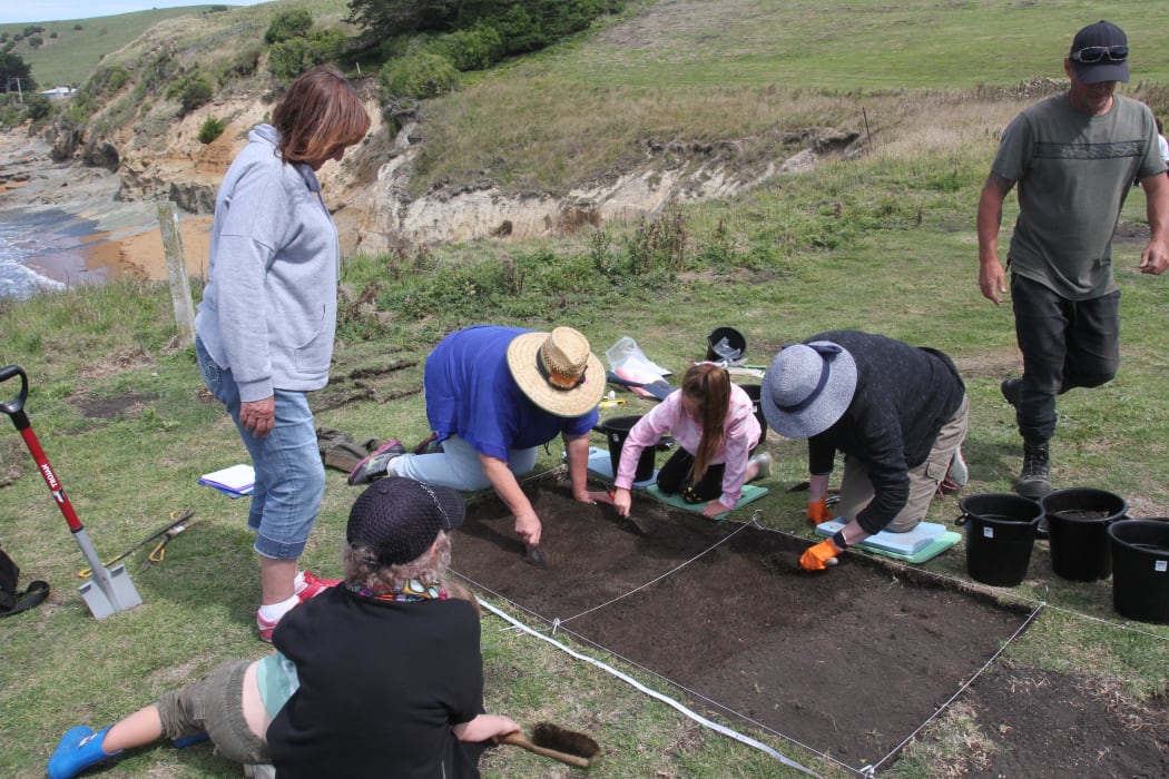 Whānau members take part in excavation. People are crowded around a dig site using small trowels to dig it up.