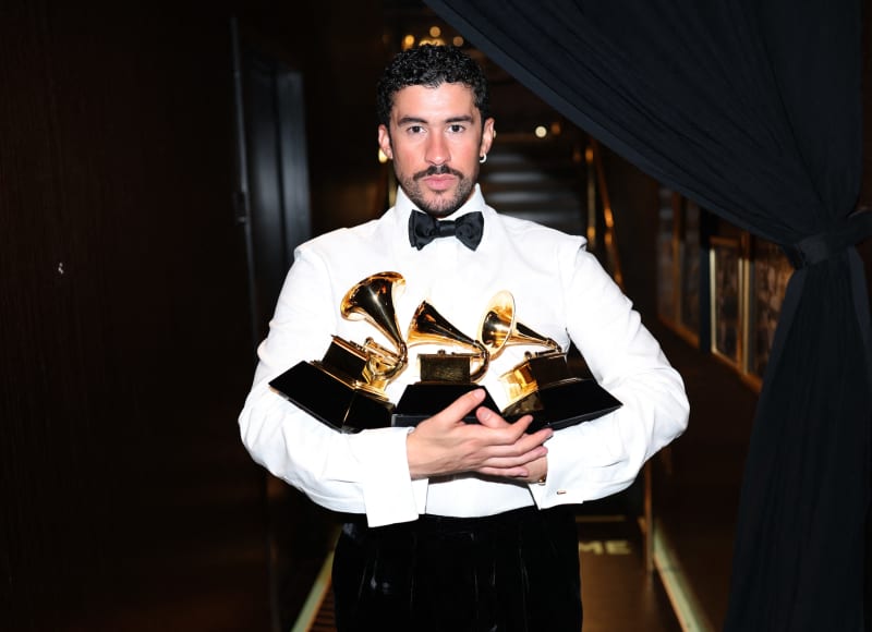 Bad Bunny, wearing a white shirt and black bowtie, bundles three golden trophies in his arms.