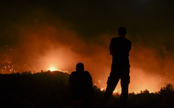 People watch the fires near the village of Malona in the Greek island of Rhodes on July 23, 2023. Tens of thousands of people fled wildfires on the Greek island of Rhodes on July 23, 2023, as terrified tourists scrambled to get home. Firefighters tackled blazes that erupted in peak tourism season, sparking the country's largest-ever wildfire evacuation -- and leaving flights and holidays cancelled. (Photo by SPYROS BAKALIS / AFP)