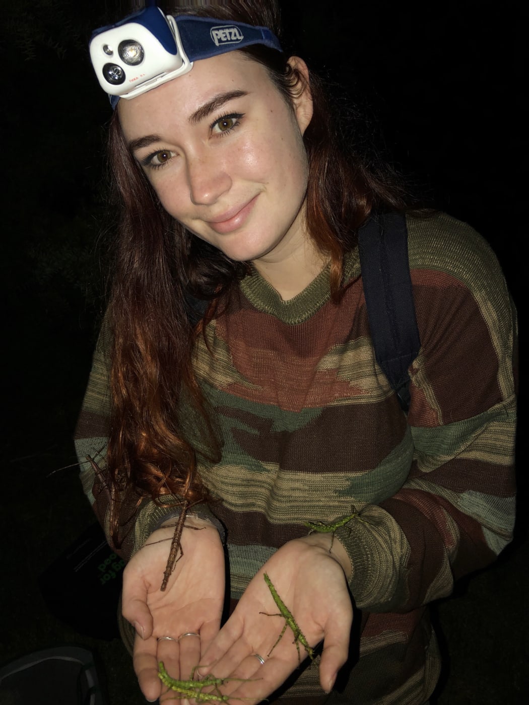 Morgane smiles at the camera. It is night time. She is wearing a headtorch and holding stick insects in her hands.