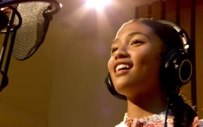 Jaya is in a recording studio, wearing headphones and looking towards the microphone. She is smiling.