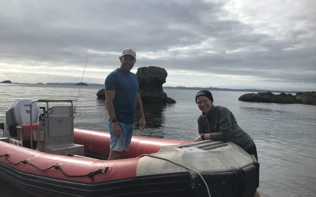 A man stands in an inflatable zodiac boat pulled ashore in the shallows while a woman stands next to the boat. The pair are looking at the camera and smiling.