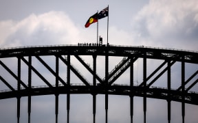 (FILES) Tourists stand under an Aboriginal and Australian national flag located on the top of the Sydney Harbour Bridge on September 27, 2023, ahead of the upcoming "Voice" referendum. A referendum aimed at elevating the rights of Indigenous Australians has instead triggered a torrent of racist slurs and abuse, with toxic debate spreading online and in the media. The October 14 vote will decide whether to finally recognise First Nation peoples in the constitution as Australia's first inhabitants. (Photo by DAVID GRAY / AFP)