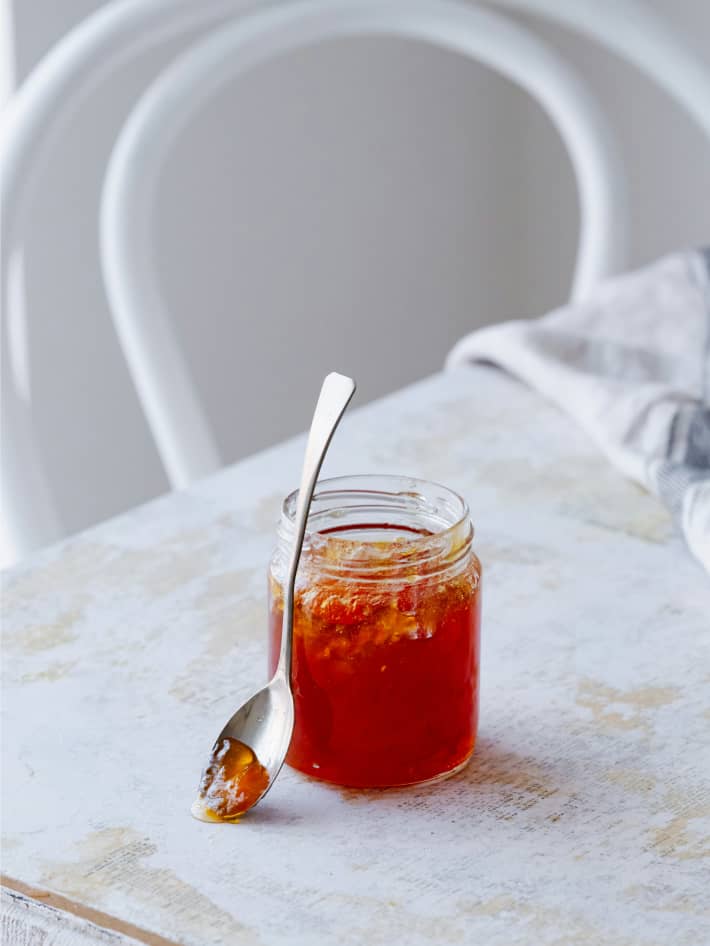 A jar of tomato chilli jam on a white table with a spoon propped against the jar,