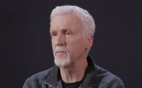James Cameron has white hair and a white goatee and wears a grey sweater.