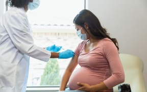 Wearing protective masks because of COVID-19, a mid adult pregnant woman watches as the female technician administers a booster shot.