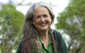 Wendyl smiles at the camera. She is outside in a clearing surrounded by lush bush. The sea is in the distance. It is a bright, pale day.
