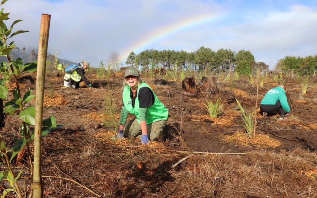A woman smiling broadly while kneeling next to seedlings in a planting site. She is wearing bright green with a green cap and gardening gloves. There are two people kneeling in the background behind her, planting trees, and a rainbow stretches across the cloudy sky.