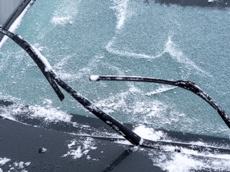 Windshield of car is covered with light snow and wipers are raised from glass.