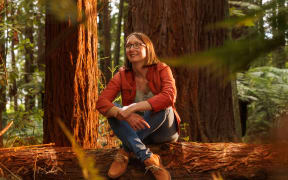 Hilary Corkran is sitting cross-legged on a fallen pine log in a forest. She is smiling and wearing tan leather boots.