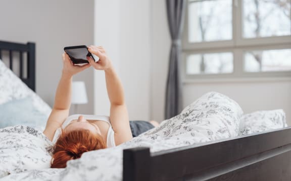 Young woman spending a relaxing day at home lying on her back on the bed reading a message on her mobile phone