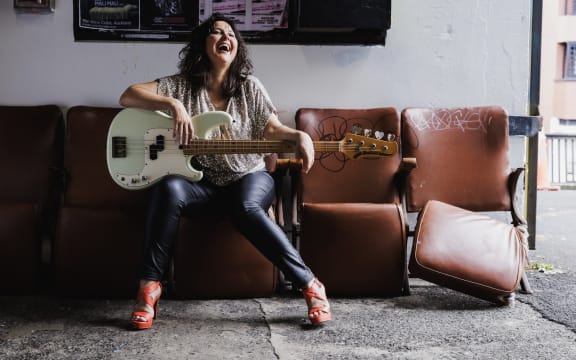 Jodie Rimmer is in character as Nichola Cheesman. She's sitting on a brown lather chair and holdin a bass guitar. She's looking up to the left and laughing.