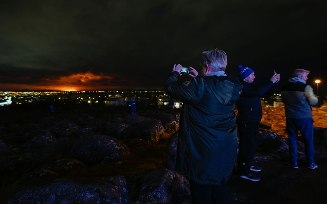 People on the outskirts of Reykjavik take pictures and selfies with the orange coloured sky as molten lava flows out from a fissure on the Reykjanes peninsula north of the evacuated town of Grindavik, western Iceland on March 16, 2024. Lava spewed Saturday from a new volcanic fissure on Iceland's Reykjanes peninsula, the fourth eruption to hit the area since December, authorities said. (Photo by Halldor KOLBEINS / AFP)