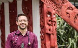 Dr Mataroria Lyndon, Senior Lecturer in Medical Education at the University of Auckland, stands in front of a wharenui wearing a button-up shirt smiling.