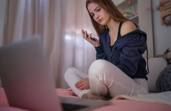 Young girl looks at her laptop on her bed as she holds up her phone.