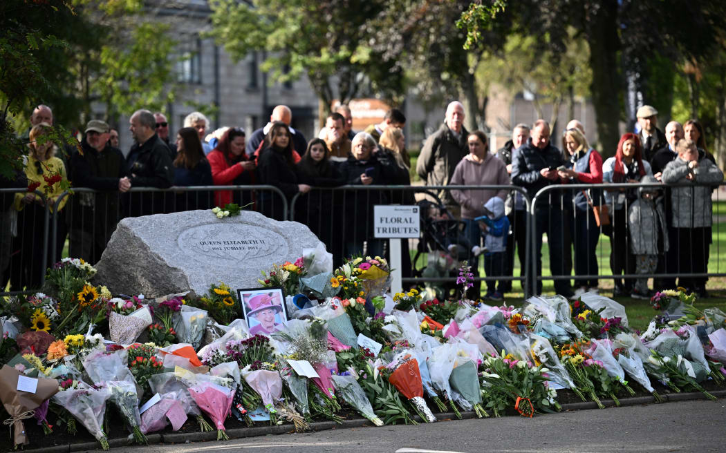 Members of the Public gather by a makeshift memorial to pay respects as they wait for the hearse carrying the coffin of Queen Elizabeth II to pass through Ballater, on September 11, 2022. - Queen Elizabeth II's coffin will travel by road through Scottish towns and villages on Sunday as it begins its final journey from her beloved Scottish retreat of Balmoral. The Queen, who died on September 8, will be taken to the Palace of Holyrood House before lying at rest in St Giles' Cathedral, before travelling onwards to London for her funeral. (Photo by Paul ELLIS / AFP)