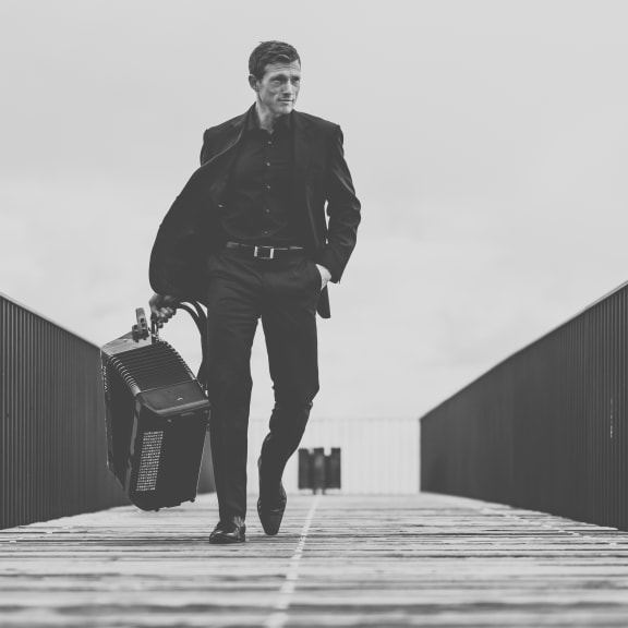 A man walks across a walkway, holding an accordion in his right hand. His left hand is in his pocket.