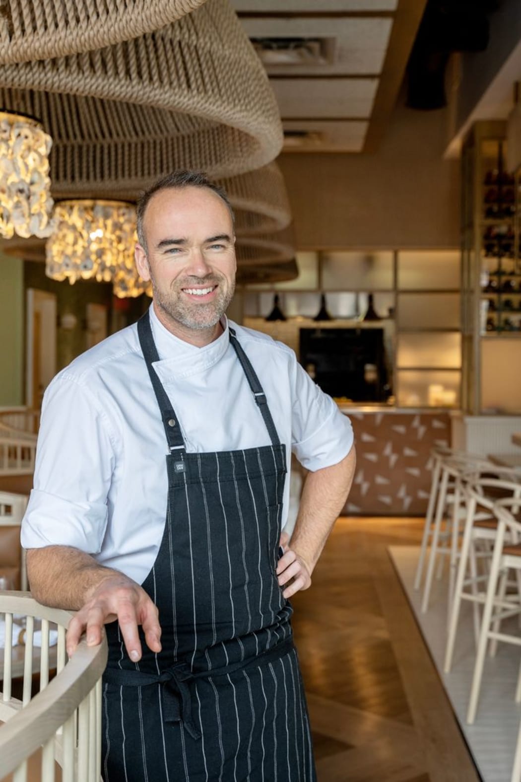 Toby stands in his restaurant wearing an apron over a white chef's shirt. He smiles.