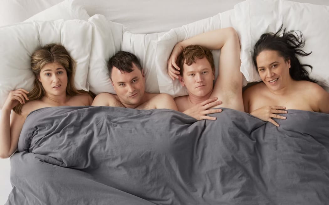 Everyone is nude in bed, covered by a grey duvet.