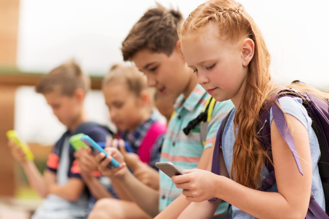 primary education, friendship, childhood, technology and people concept - group of happy elementary school students with smartphones and backpacks sitting on bench outdoors